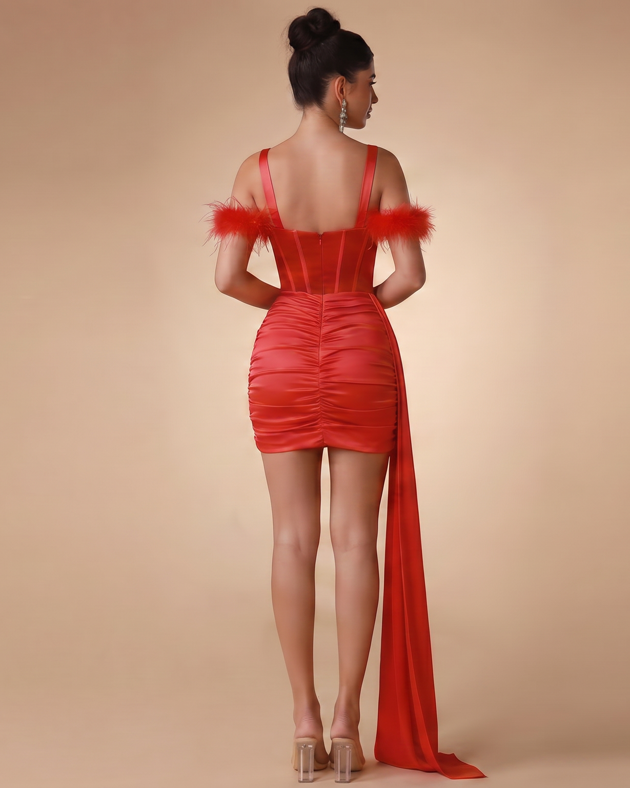 A woman stands with her back to the camera wearing a bright red off-the-shoulder mini dress with a long draped train, photographed against a plain warm beige studio background.