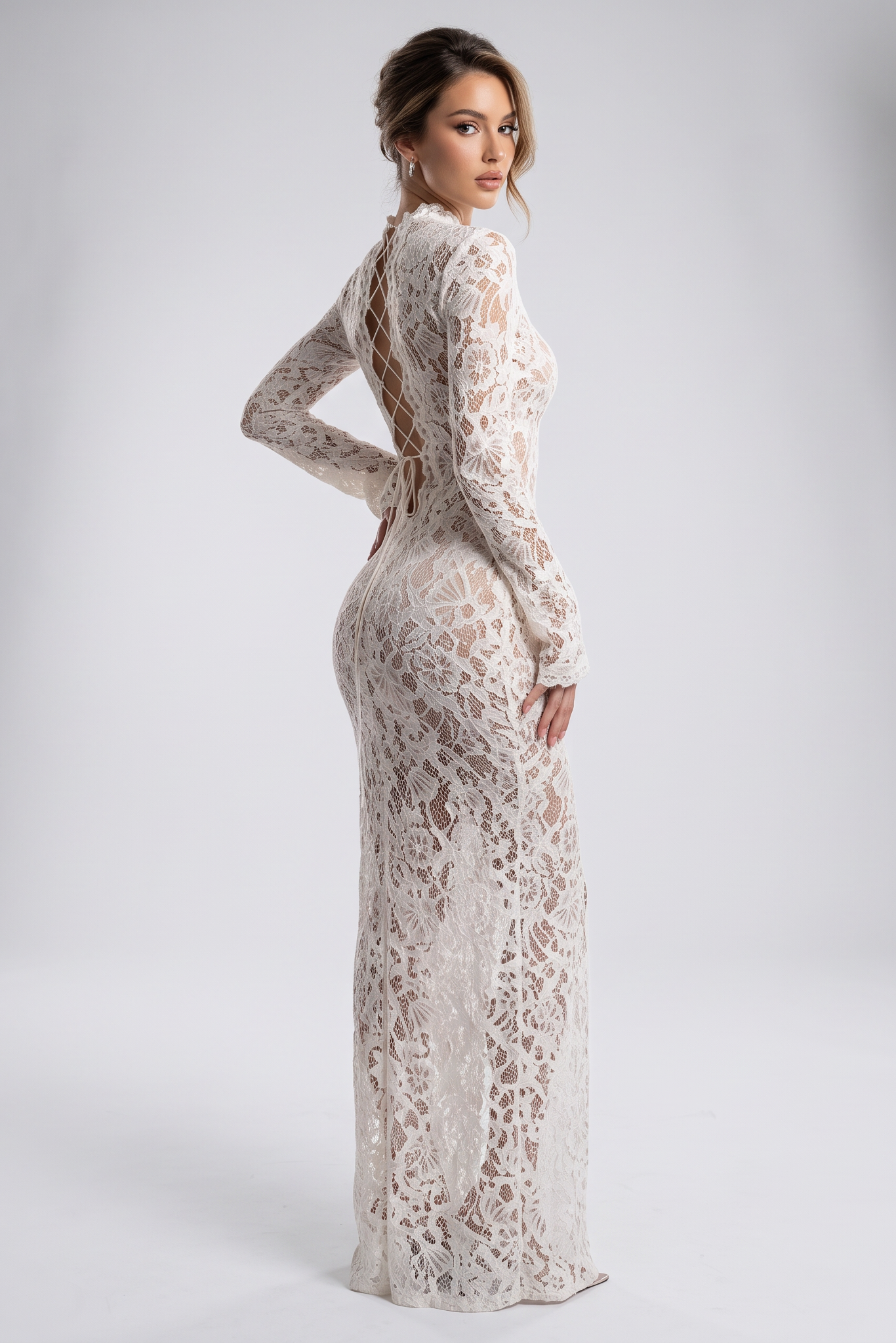 A woman stands in a studio wearing a long-sleeve lace maxi dress with an open back. Studio lights are visible on both sides against a light gray backdrop.