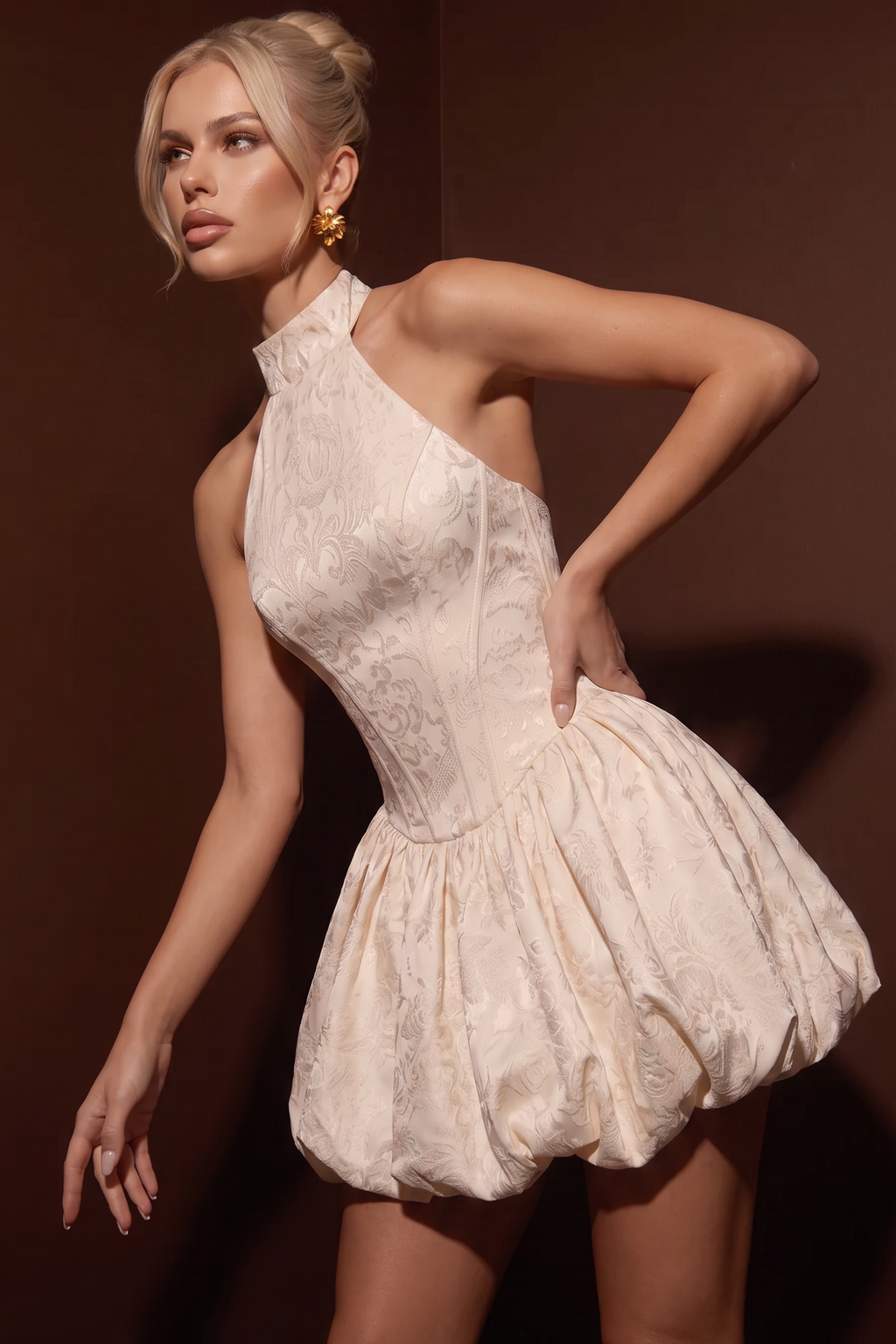 A woman modeling a sleeveless, high-neck, one-shoulder mini dress with a structured corset-style bodice and voluminous gathered skirt, in a light champagne or cream tone against a dark brown studio background.