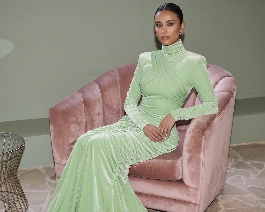 A woman wearing a light mint green velvet long-sleeve turtleneck gown sits on a dusty pink velvet armchair in a styled interior setting.